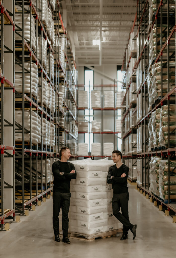 Two men standing in a warehouse with a large stack of bags.
